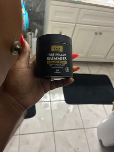 Customer's hand holding a black jar of XARA SHILAJIT Pure Shilajit Gummies in a bathroom.