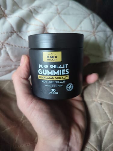 Reviewer holding a black jar of XARA SHILAJIT Pure Shilajit Gummies against a quilted background.
